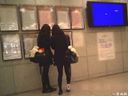 A pair of J flapping their legs in black tights in front of the timetable